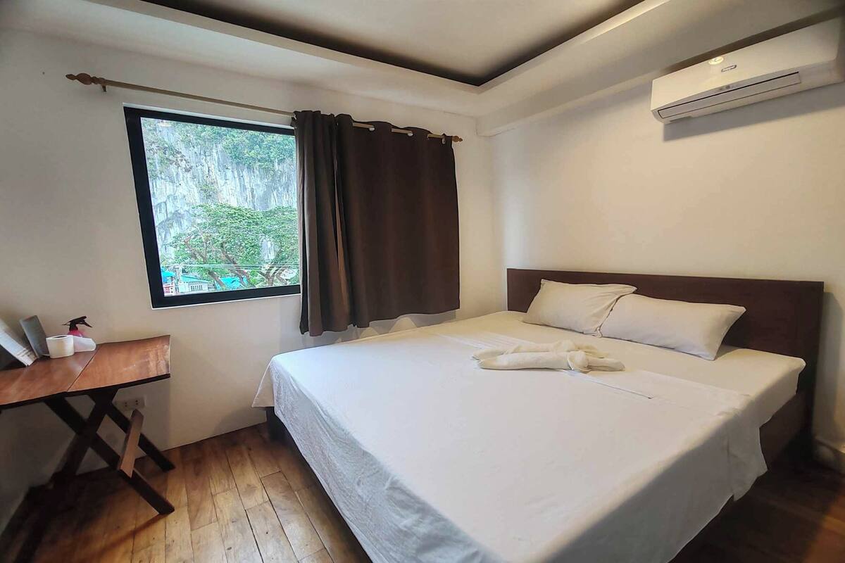 A bedroom with a bed, a desk, a window with curtains, and an air conditioning unit.