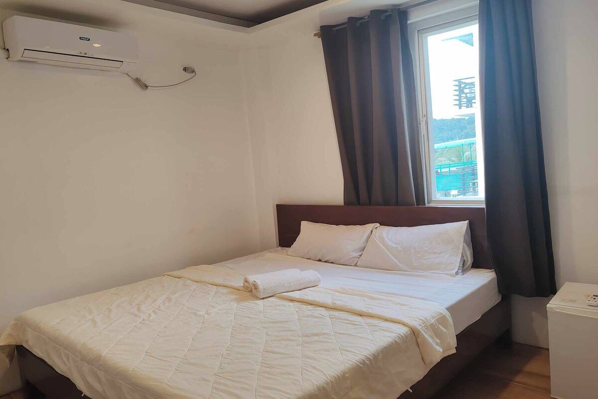 A neatly made bed with a white duvet and pillows, a window with dark curtains, and a wall-mounted air conditioner.