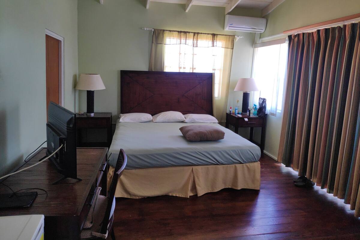 A bedroom with a wooden bed, two bedside tables with lamps, a window with curtains, and an air conditioning unit.