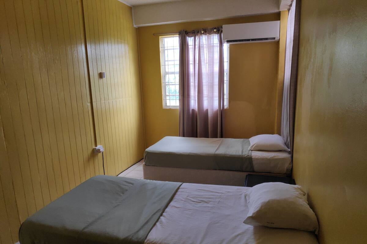 A small, compact room with two single beds, a window with curtains, and a wall-mounted air conditioning unit.