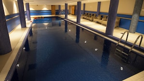 An indoor swimming pool with a tiled floor, a pool ladder, and seating areas along the sides.