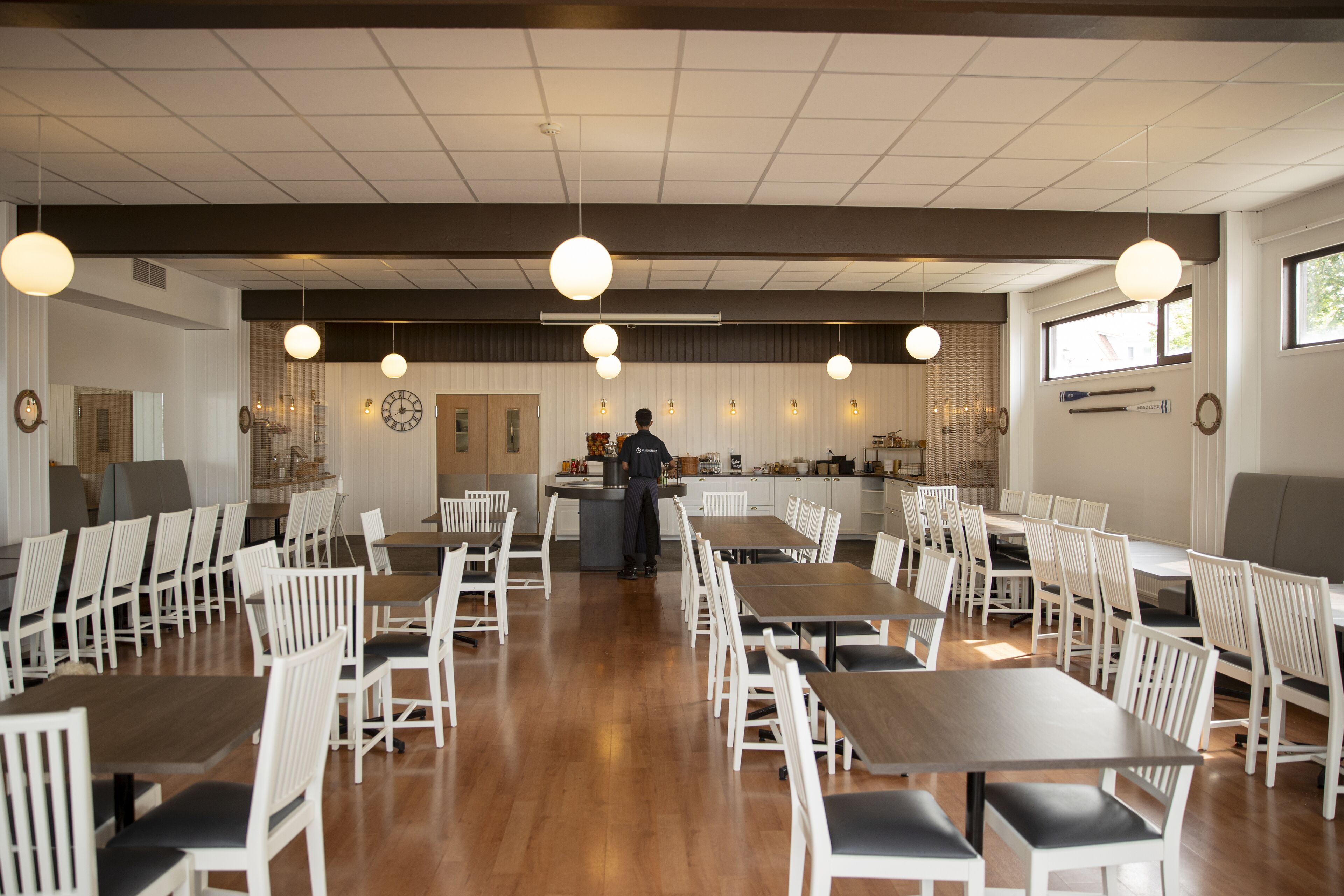 A spacious dining area with multiple tables and white chairs, a kitchen in the background, and pendant lighting.
