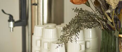 A kitchen counter with stacked white mugs, a stainless steel coffee maker, and a vase with dried flowers.