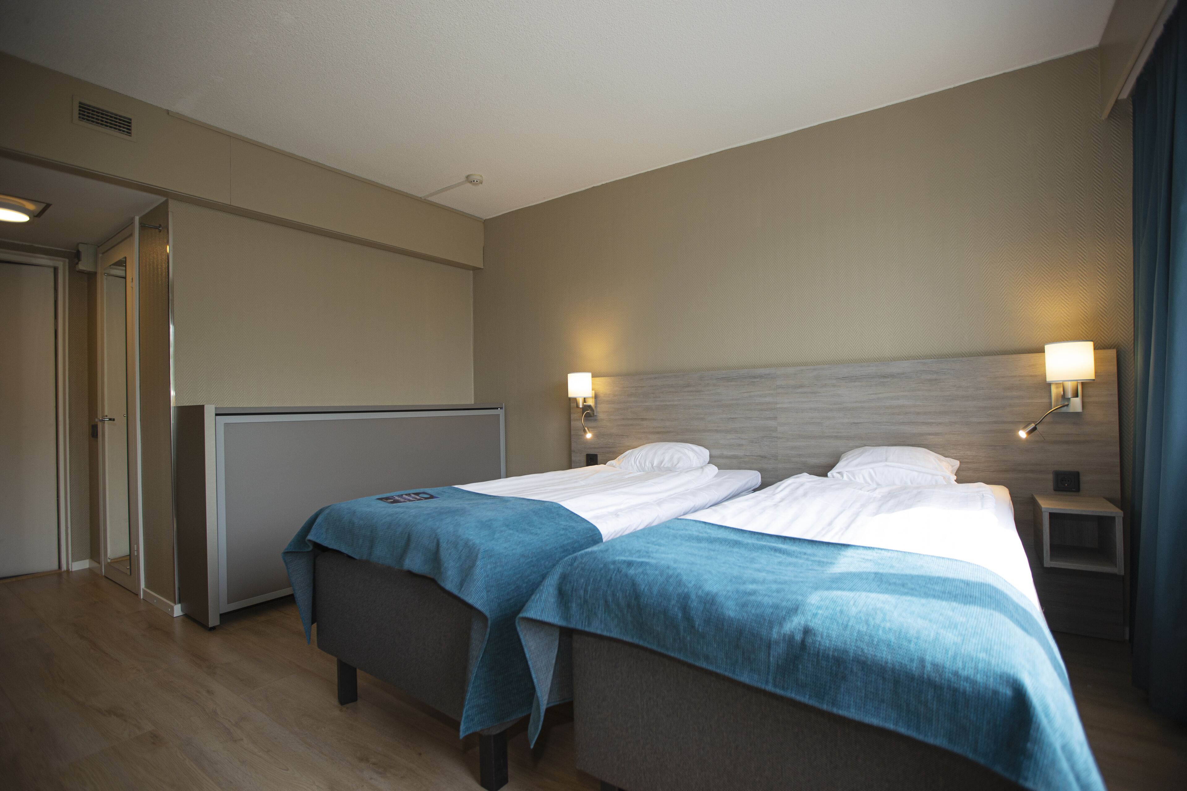 A hotel room with two beds, a bedside table with a lamp, a wardrobe, and a door leading to another room.