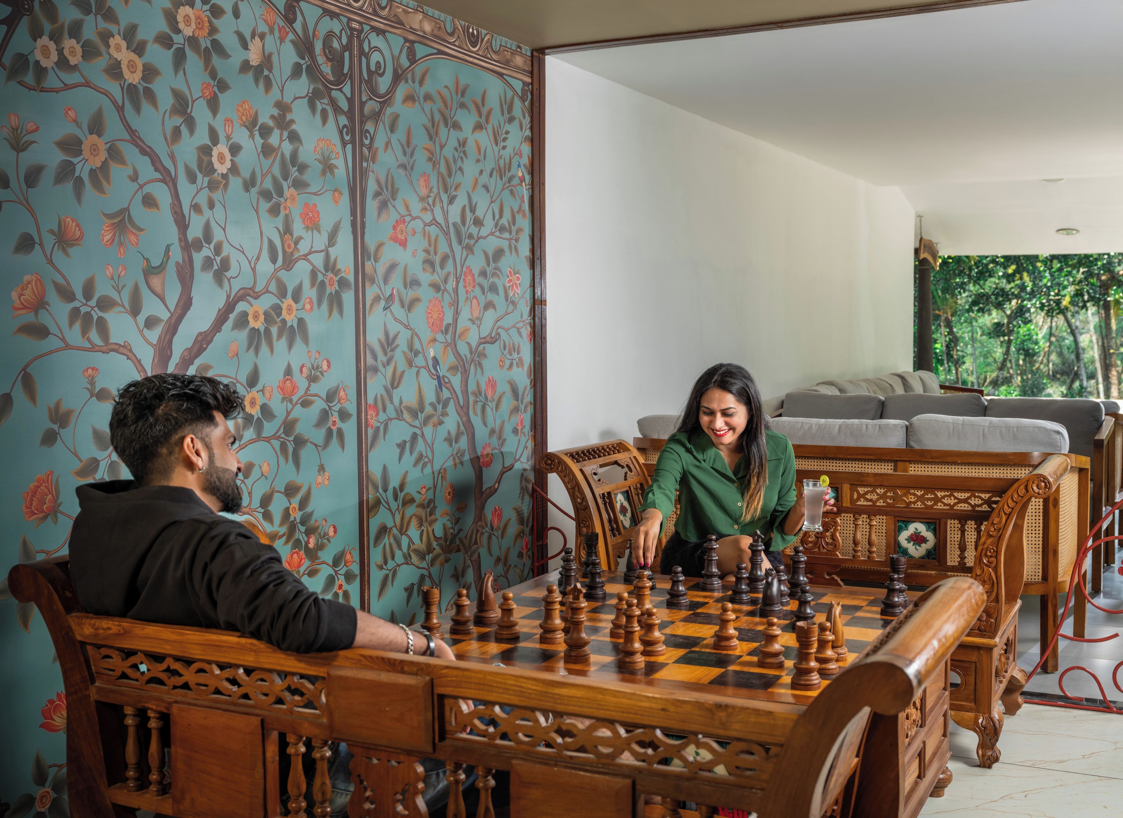A chessboard set on a wooden table in a room with floral wallpaper and a sofa in the background.