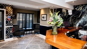 A reception area with a wooden desk, a vase of flowers, a computer, and a seating area with patterned chairs. - Thames Riviera Hotel, Sure Hotel Collection by Best Western (Maidenhead)