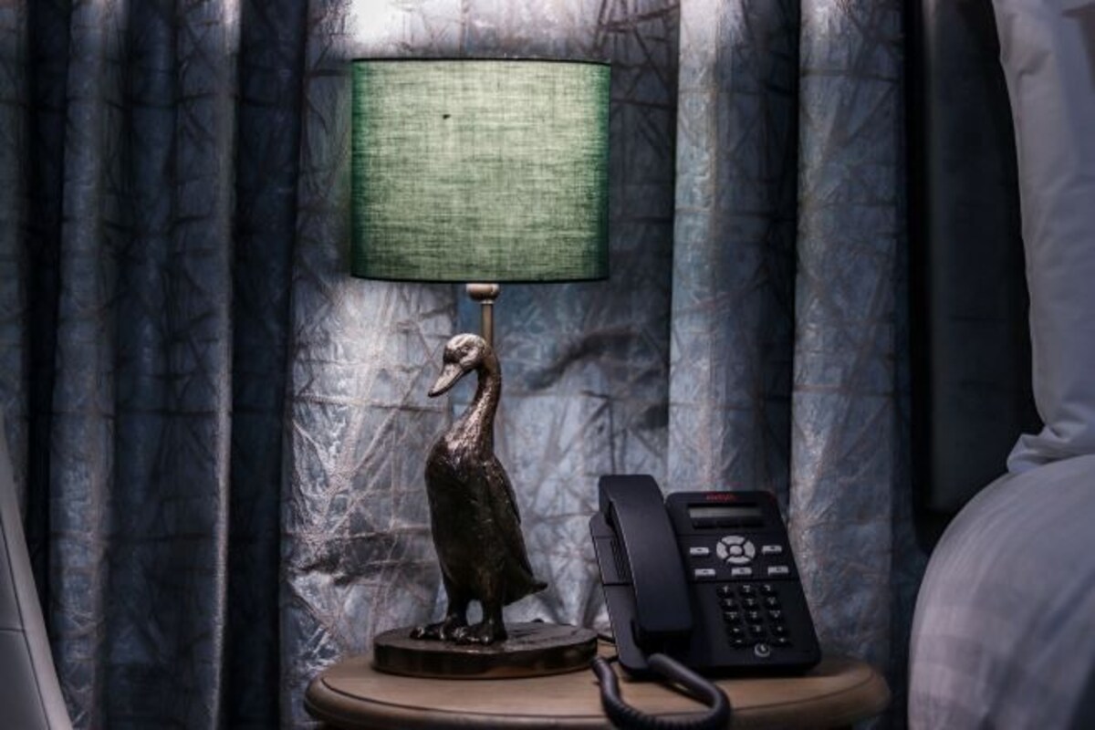 A lamp with a green shade and a bird figurine on a bedside table next to a telephone.