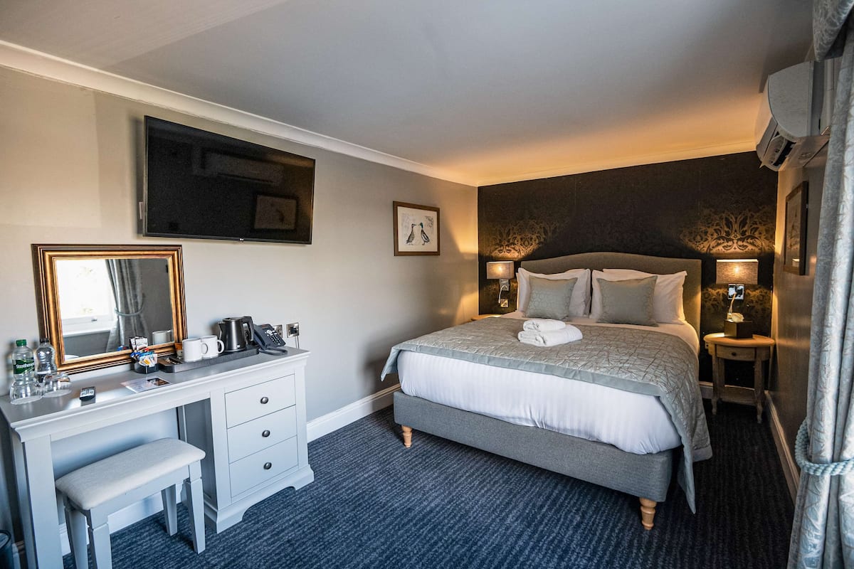 A hotel room with a bed, a desk with a mirror, a chair, a bedside table with a lamp, and a wall-mounted TV.