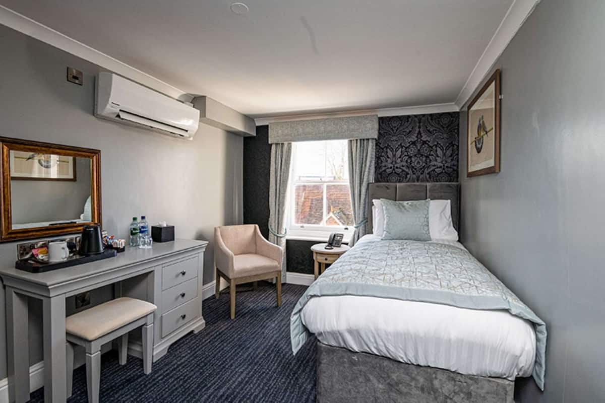 A hotel room with a bed, a desk with drawers, a chair, an air conditioner, a window with curtains, and a framed picture on the wall.