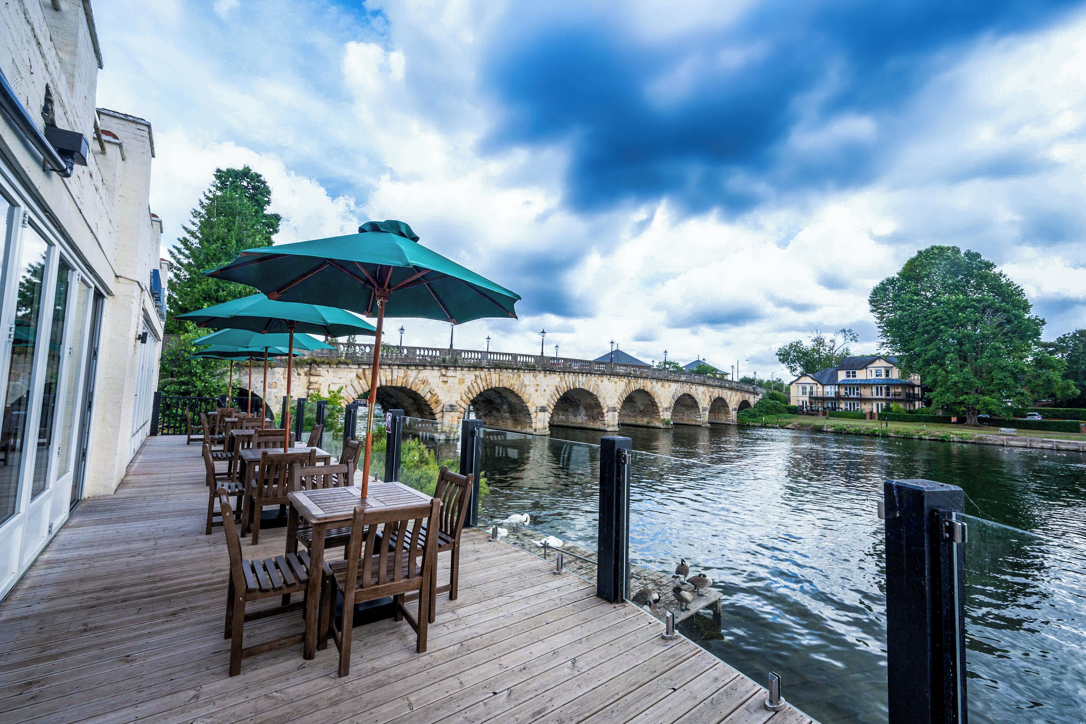 A riverside deck with wooden tables and chairs, a stone bridge in the background, and a building on the opposite bank.