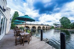 A riverside deck with wooden tables and chairs, a stone bridge in the background, and a building on the opposite bank. - Thames Riviera Hotel, Sure Hotel Collection by Best Western (Maidenhead)