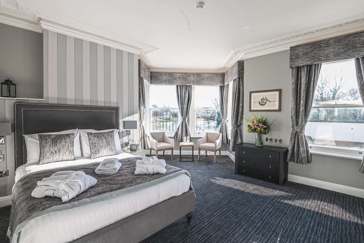 A hotel room with a large bed, two armchairs, a small table, and a view of the city through large windows.