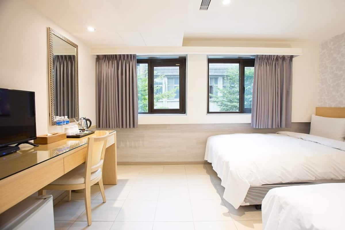 A hotel room with a bed, a desk with a chair, a television, and two windows with curtains.
