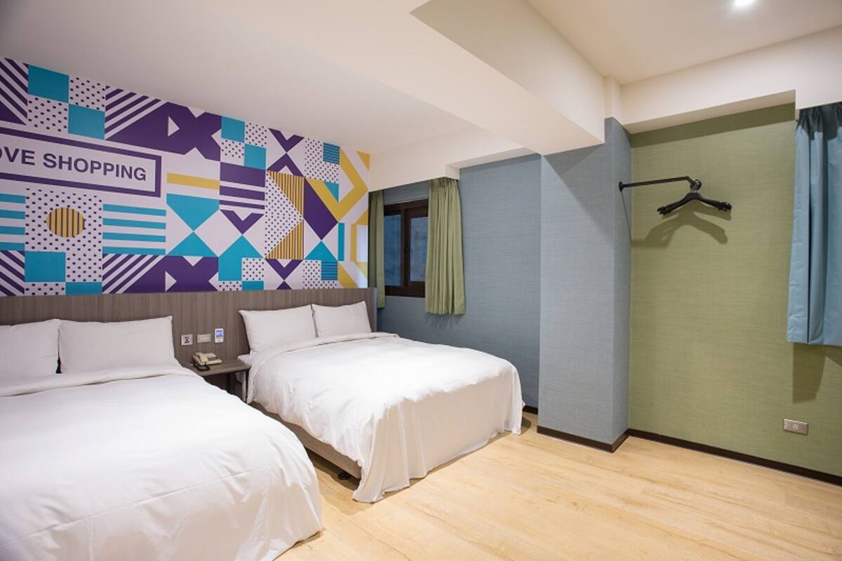 A hotel room with two beds, a geometric-patterned wall, a window with curtains, and a coat rack.