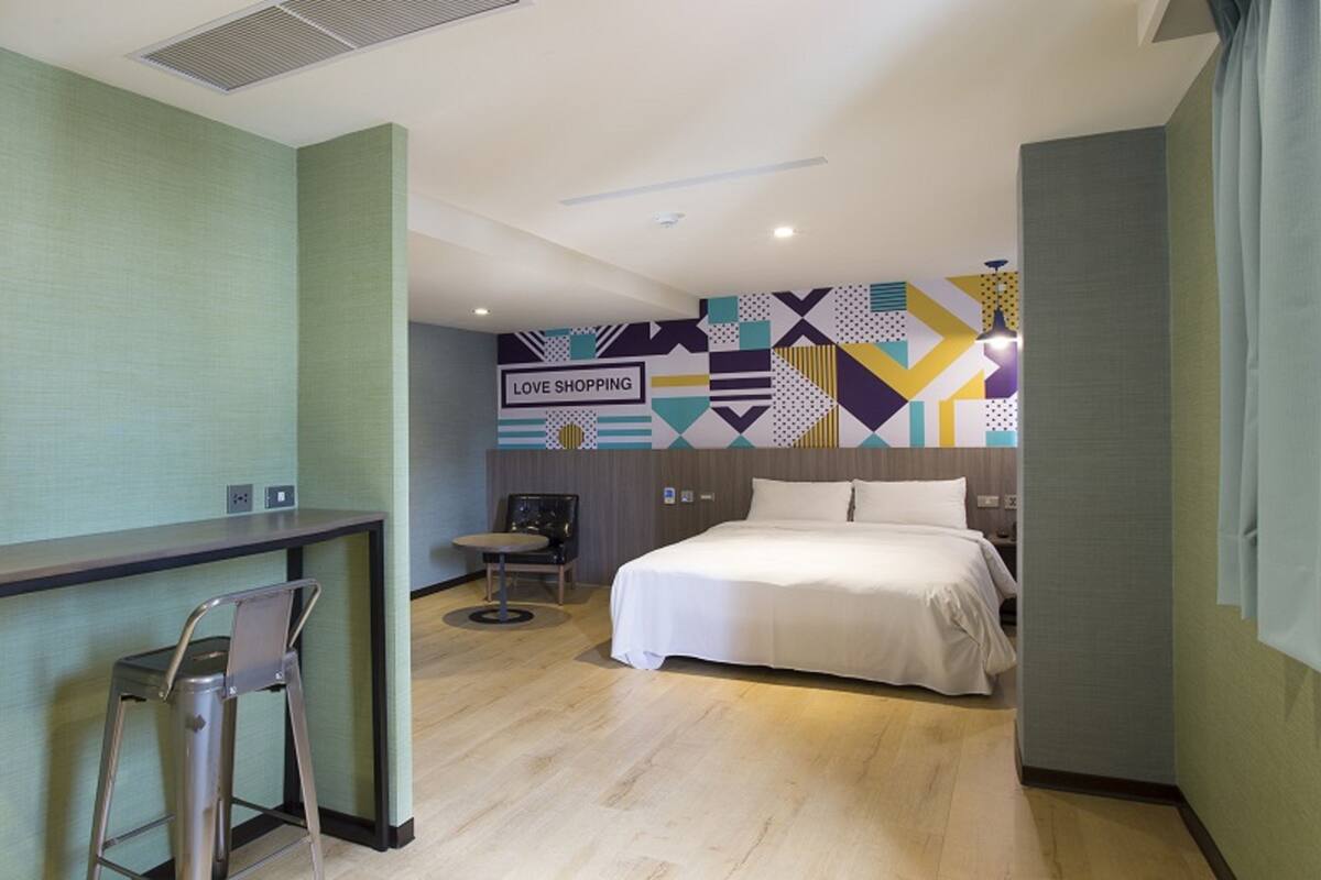 A hotel room with a bed, a desk, and a chair. There is a wall with a geometric pattern and a sign that says "LOVE SHOPPING".