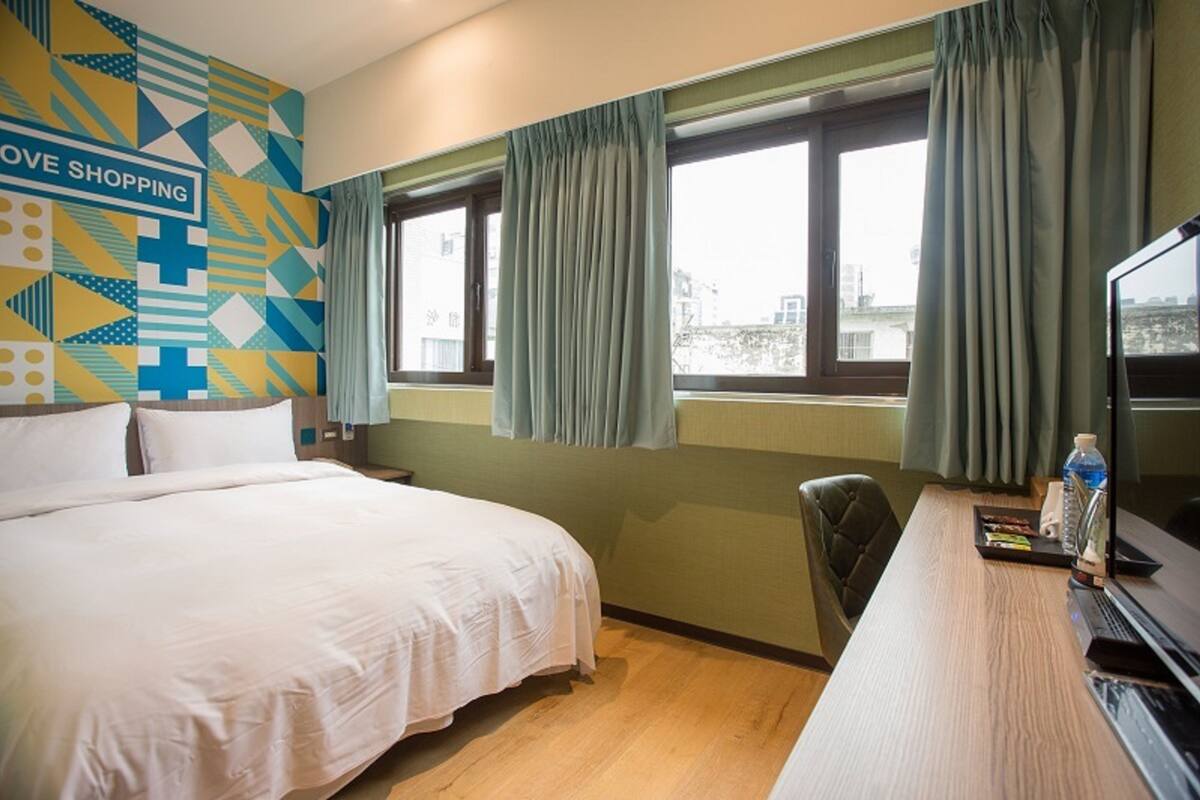 A hotel room with a bed, a desk with a computer, a chair, and a window with curtains.