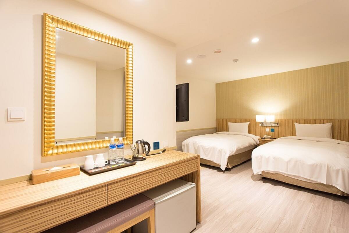 A hotel room with a large mirror, two beds, a wooden desk, a small refrigerator, and a bedside table with a lamp.