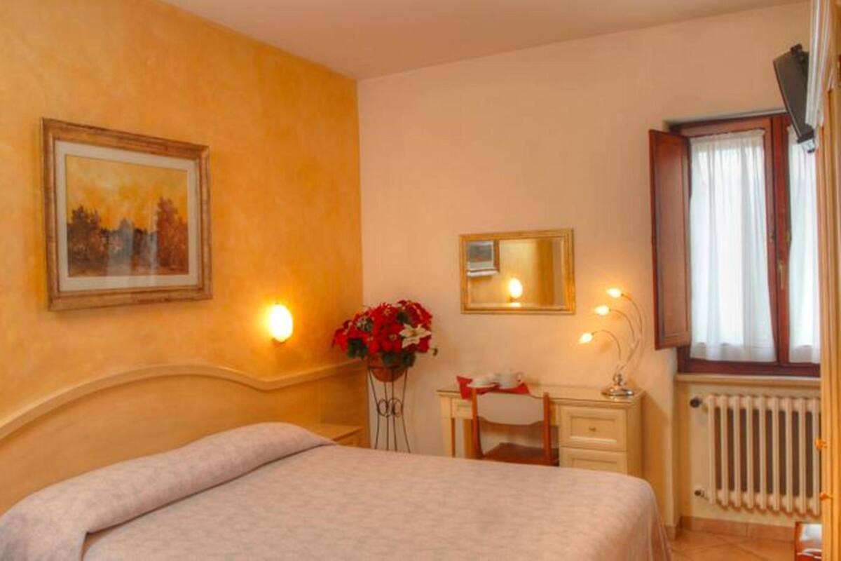 Una habitación de hotel con cama, mesita de noche, espejo, ventana con cortinas y radiador.
