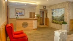 A reception area with a wooden reception desk, a red chair, a painting on the wall, and a clock above the door. - Hotel Belvedere (Roccaraso)