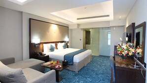 A hotel room with a bed, a desk, a chair, a sofa, a mirror, and a bathroom visible through a glass door. - Ramee Dream Resort (Seeb)