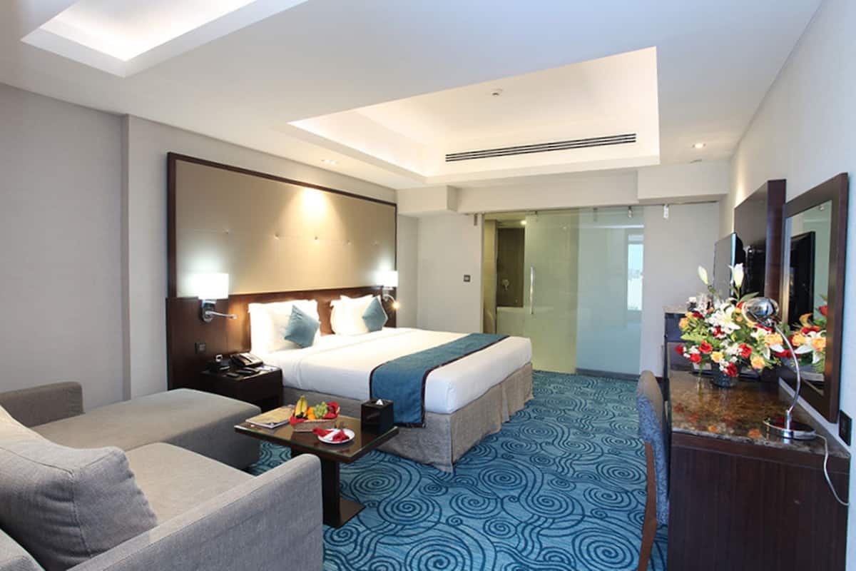 A hotel room with a bed, a desk, a chair, a sofa, a mirror, and a bathroom visible through a glass door.