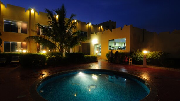 A swimming pool surrounded by a brick patio, with a building featuring multiple windows and a palm tree in the foreground. - Ramee Dream Resort (Seeb)