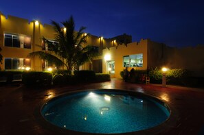 A swimming pool surrounded by a brick patio, with a building featuring multiple windows and a palm tree in the foreground.