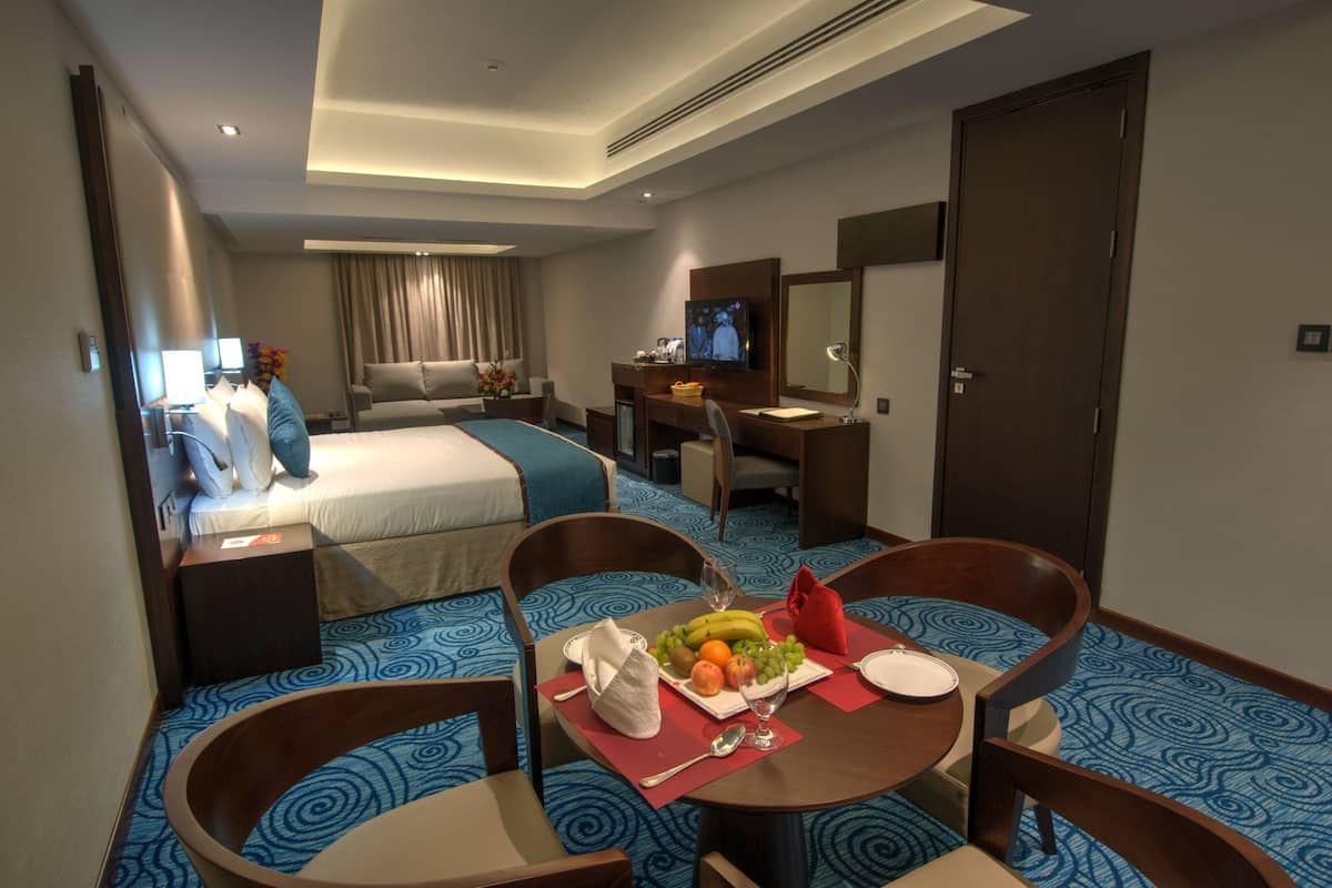 A hotel room with a bed, a desk with a TV, a dining area with a table set for two, and a bathroom visible in the background.