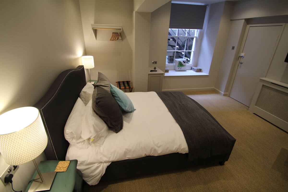 Una cama bien hecha con lámpara, una mesita de noche pequeña, una ventana con vistas y una puerta.
