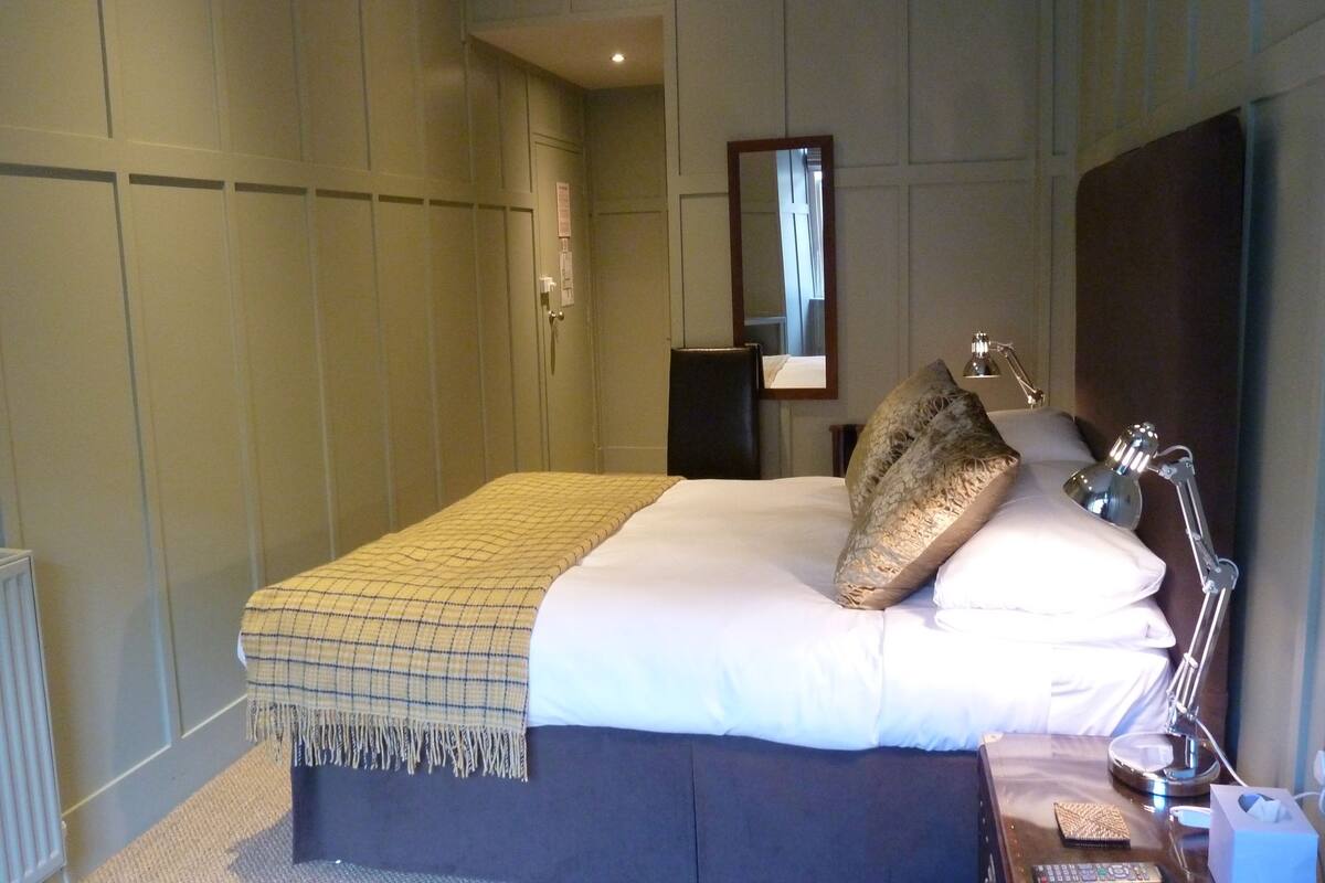 Una cama bien hecha con un plaid escocés, una lámpara de noche y una mesita de noche con un pañuelero y un control remoto.