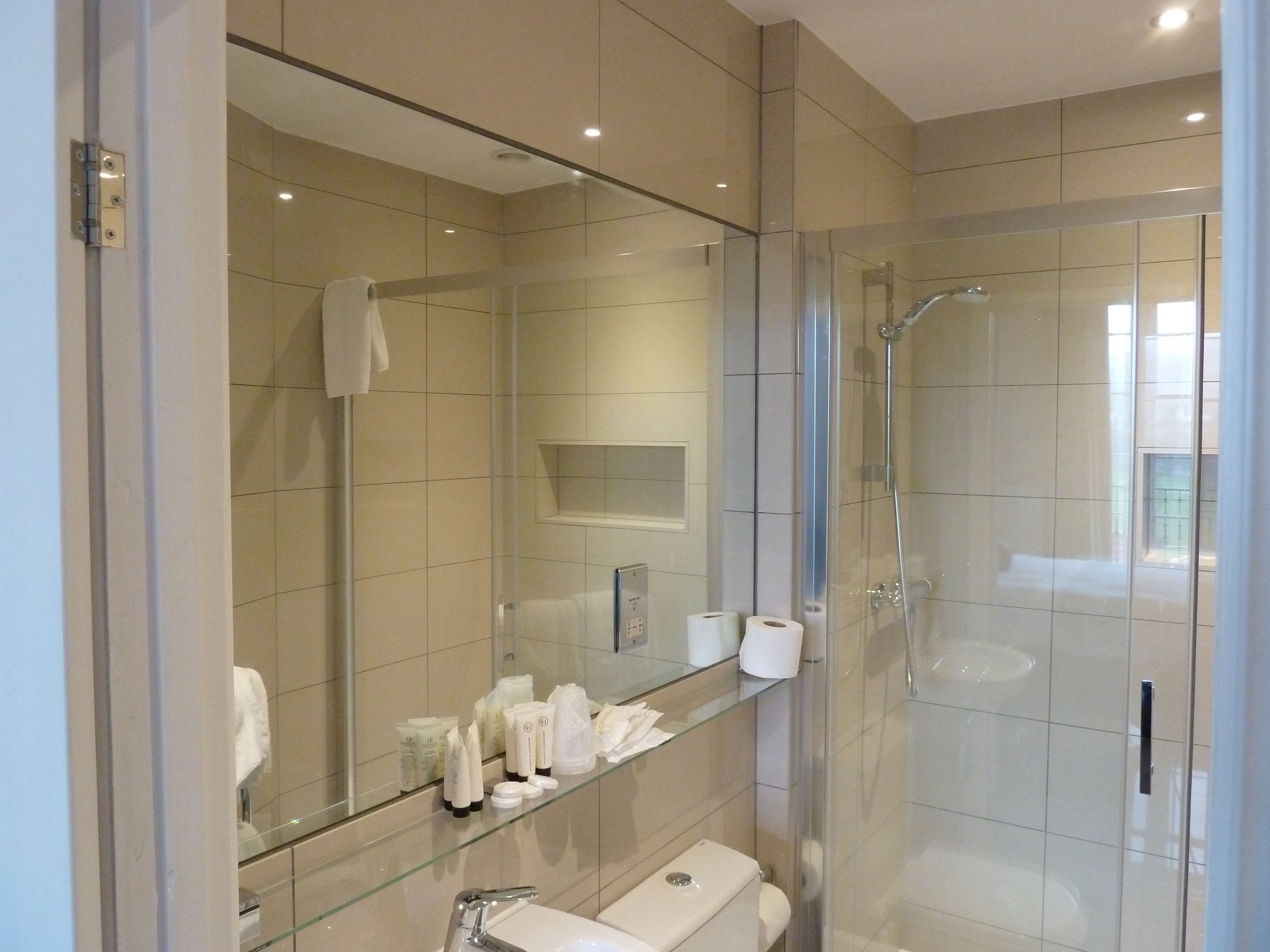 A modern bathroom with a glass shower enclosure, a large mirror, and a glass shelf with toiletries.