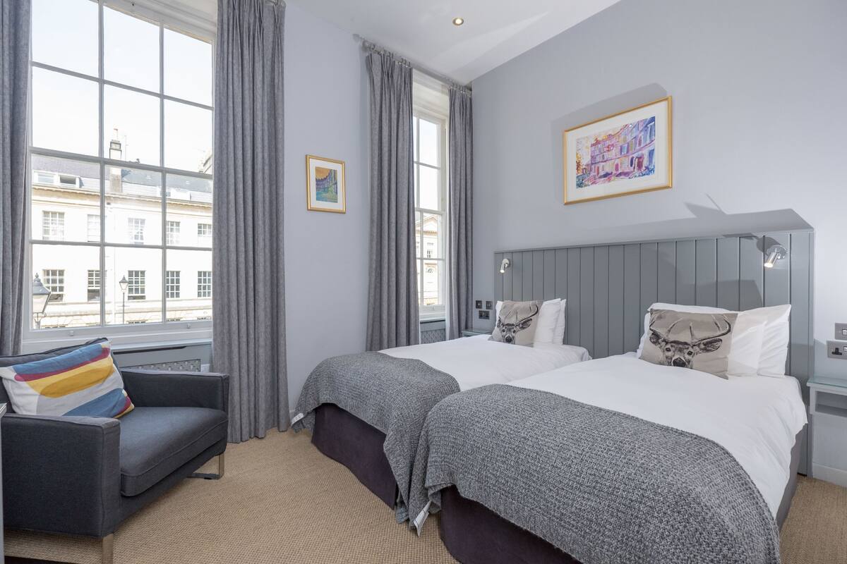Habitación de hotel con dos camas, un sofá gris, una ventana con cortinas y un cuadro en la pared.