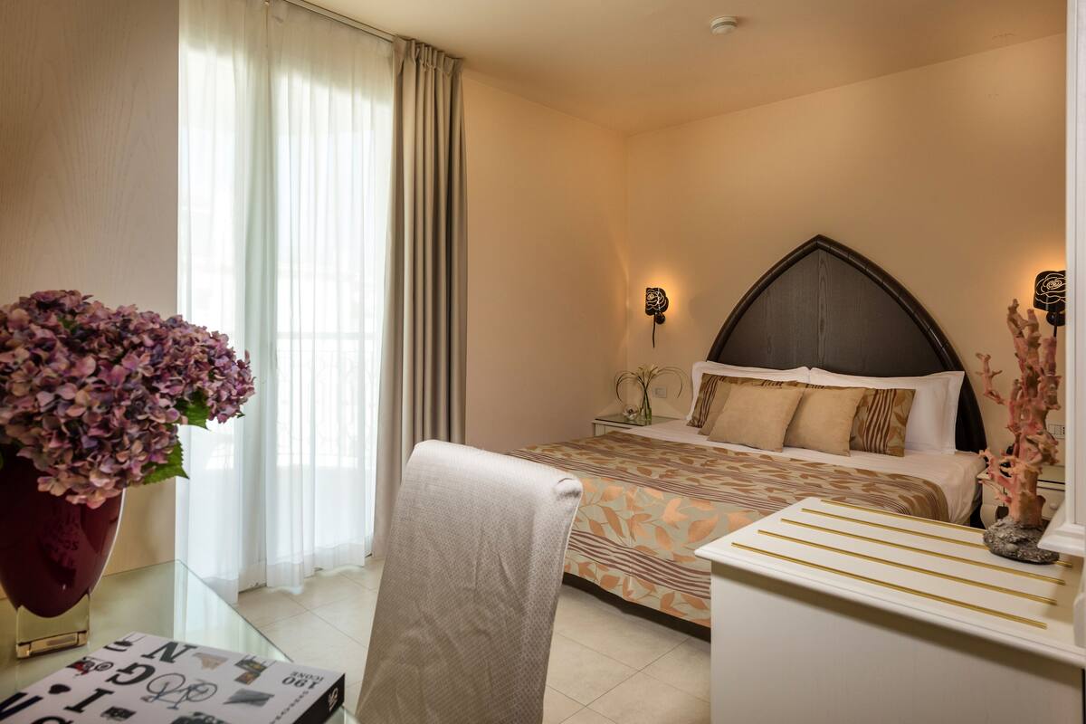 A hotel room with a bed, a bedside table with a vase of flowers, a chair, and a glass table with a magazine.