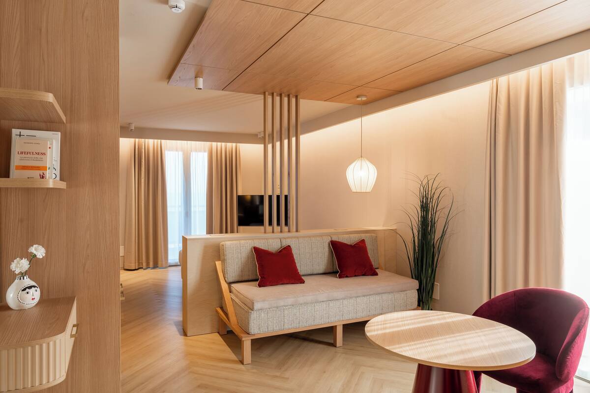 A modern hotel room with a wooden floor, a sofa with red cushions, a round table, and a tall plant.