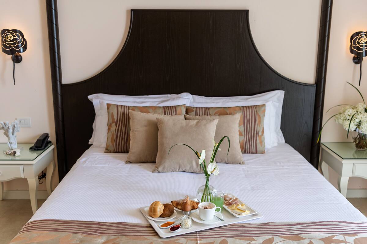 A neatly made bed with a dark headboard, white bedding, and a tray of breakfast items including pastries, a cup of coffee, and a glass of juice.
