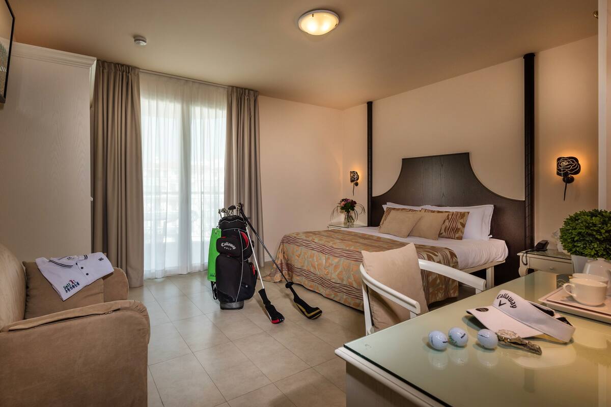 A hotel room with a bed, a sofa, a dining table, a vacuum cleaner, and a golf bag.
