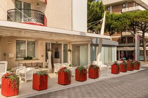 A hotel exterior with outdoor seating, large windows, and red planters with greenery. - Baia del Mar Beach Boutique Hotel (Jesolo)