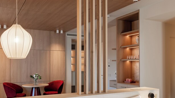 A modern hotel room with a wooden ceiling, a white bed with red pillows, a dining area with red chairs, and a built-in shelving unit with glassware. - Baia del Mar Beach Boutique Hotel (Jesolo)