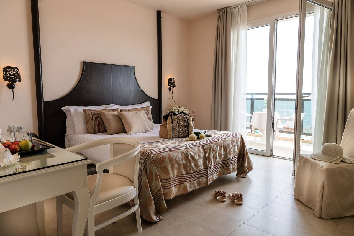 A hotel room with a bed, a desk with a tray of fruit, a chair, and a view of the balcony.