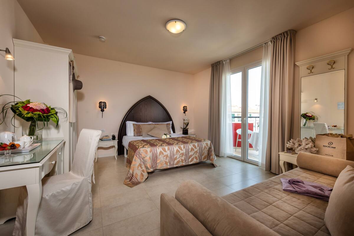 A hotel room with a bed, a sofa, a desk, and a chair. There is a window with curtains, a ceiling light, and a plant on the desk.
