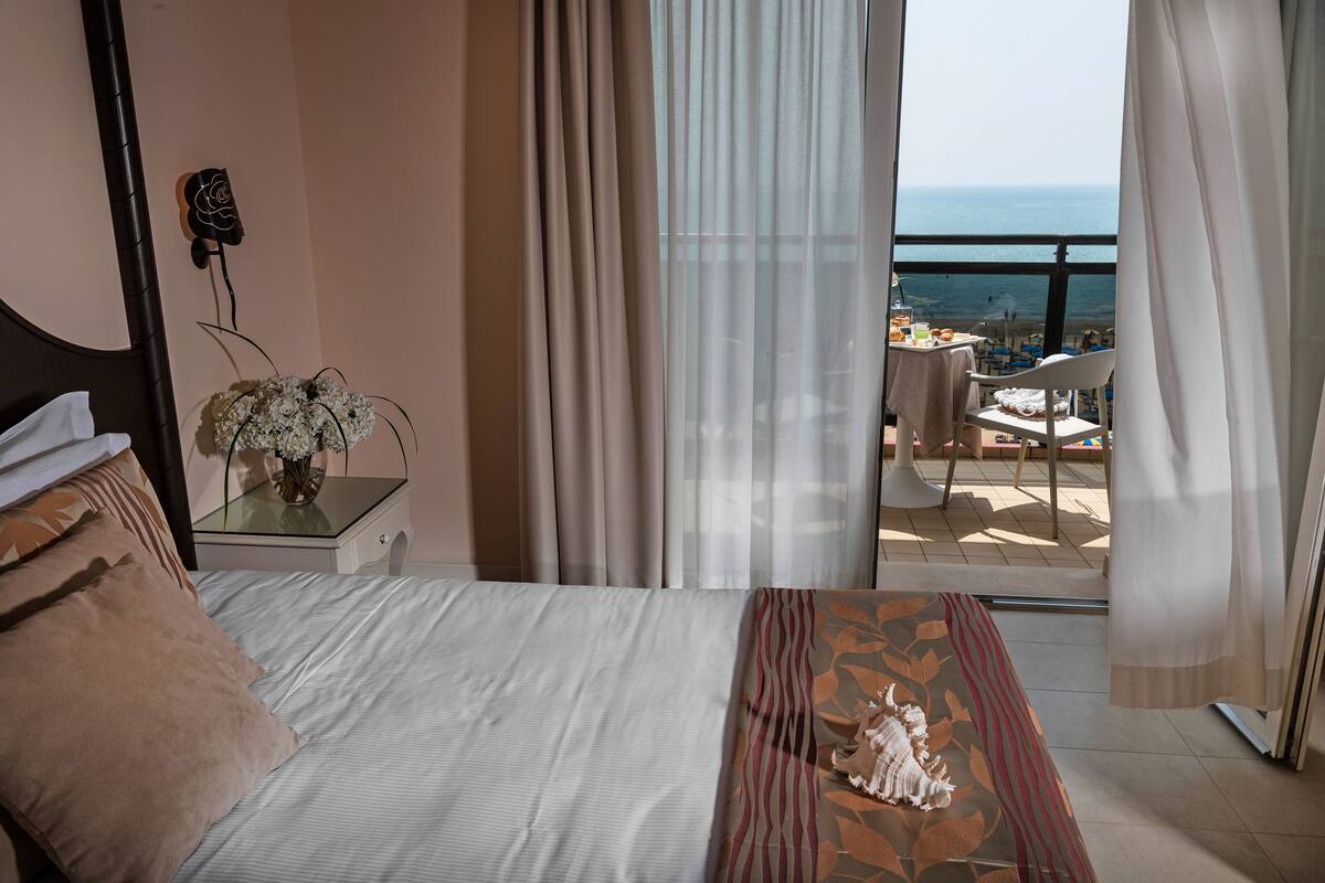 A hotel room with a bed, a nightstand with a vase, a view of the beach and ocean through sheer curtains.