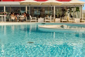 Un espace piscine agrémenté de chaises longues et de tables, entouré de baies vitrées et de plantes en pot.