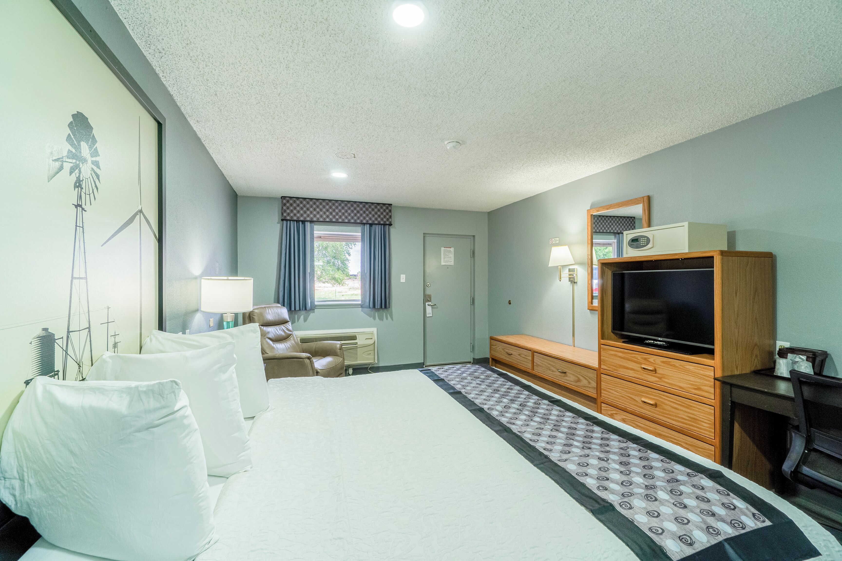 Une chambre d’hôtel comprenant un lit, une table de chevet, une lampe, une commode, une télévision et une fenêtre avec des rideaux.