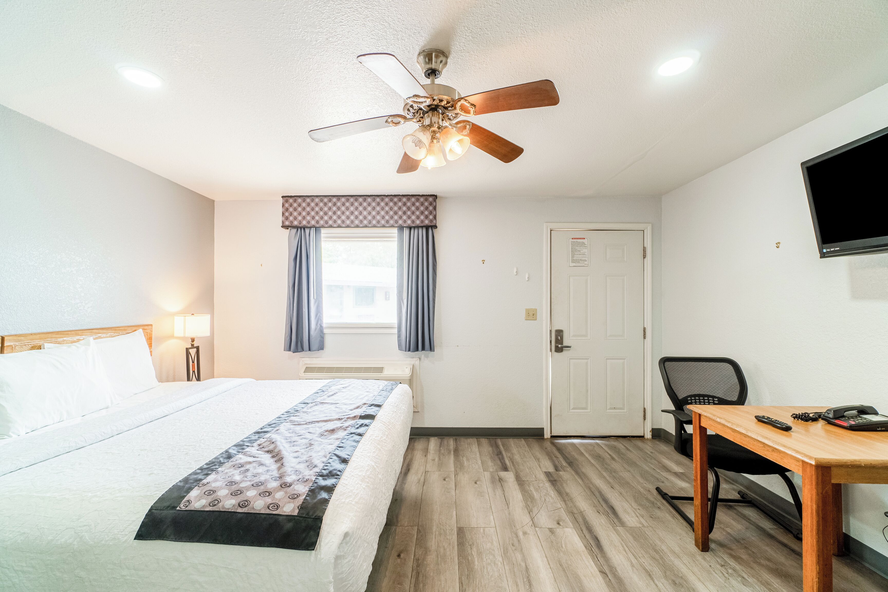 Une chambre d’hôtel équipée d’un lit, d’un bureau, d’une chaise, d’un ventilateur de plafond, d’une télévision et d’une fenêtre avec des rideaux.