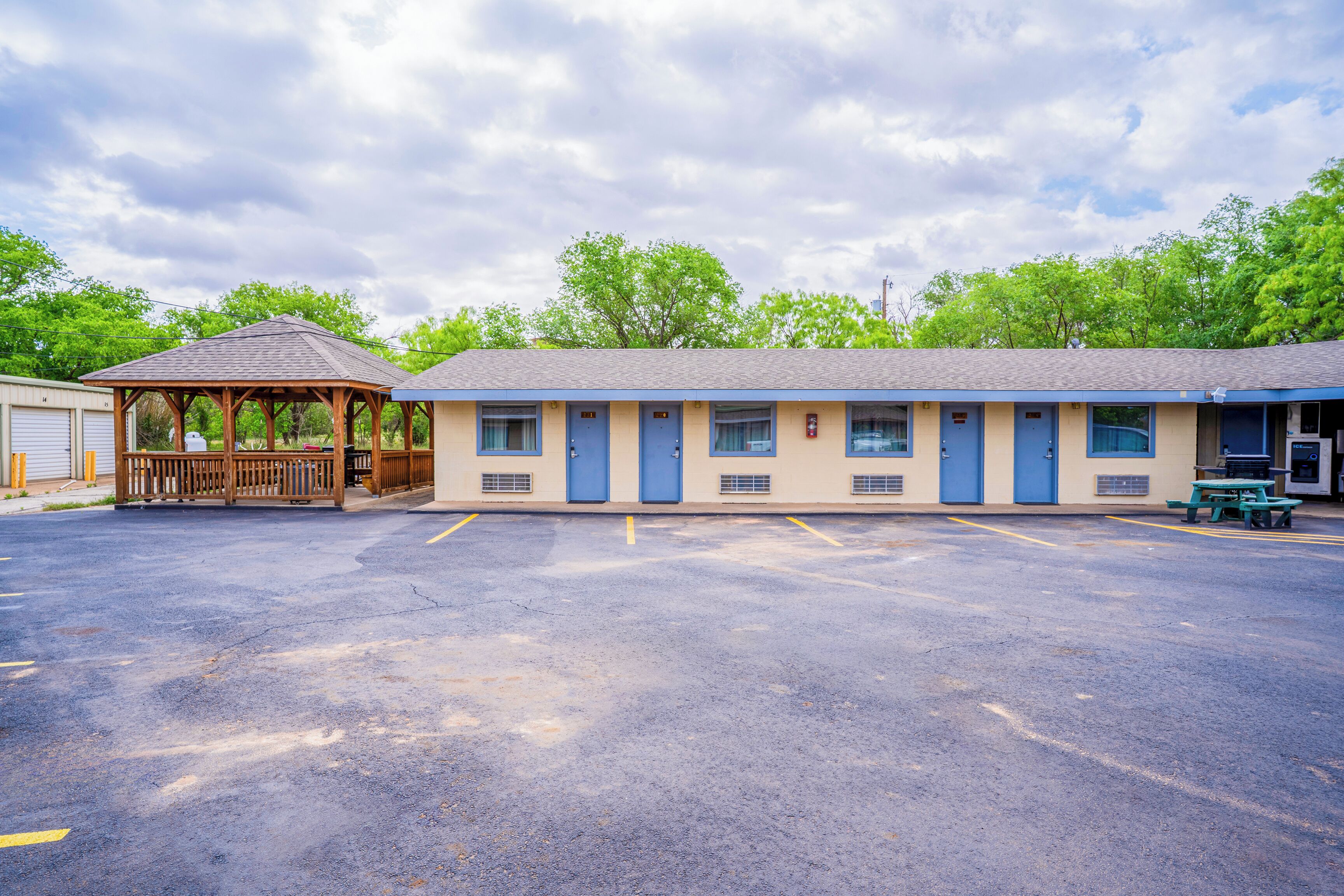 A motel with a covered outdoor seating area, a parking lot, and a building with multiple rooms.
