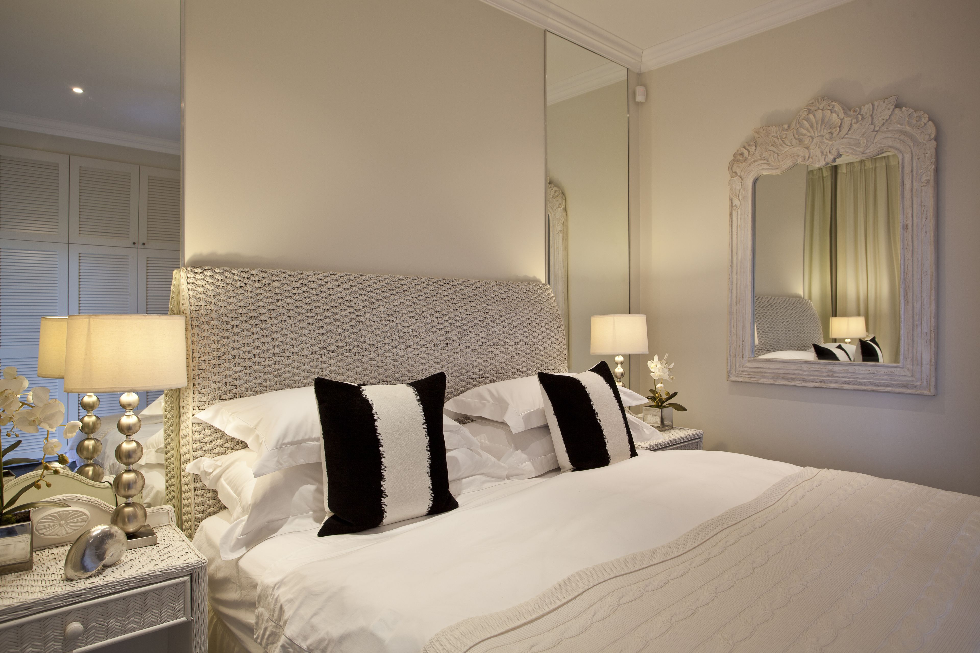 A neatly made bed with white bedding and black and white pillows, a large mirror with an ornate frame, and a bedside table with a lamp and decorative items.