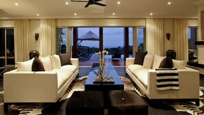 A modern living room with two white sofas, a black coffee table, and a ceiling fan. Large windows offer a view of the outdoors. - Luxury Seaside Homes (Pennington)