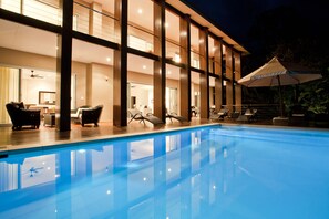 A swimming pool with lounge chairs and umbrellas, a well-lit interior with large windows, and a modern architectural design.