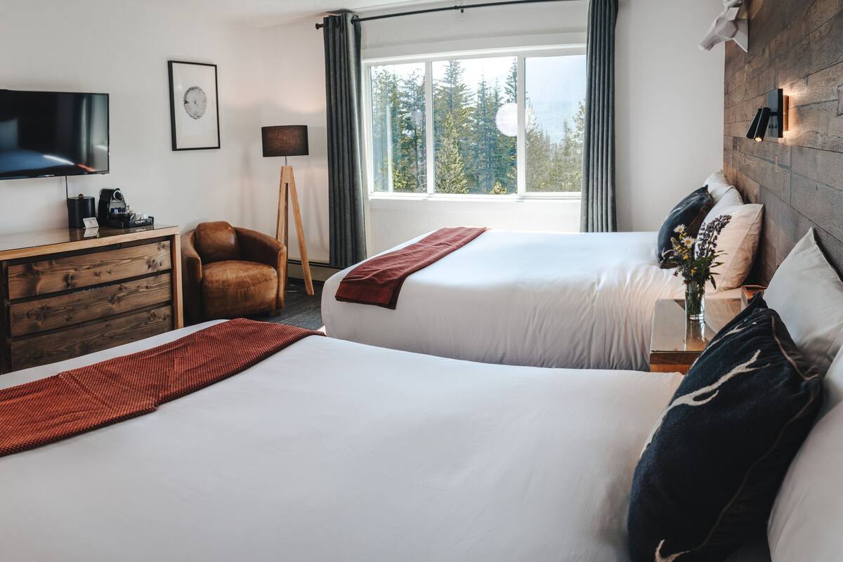 A hotel room with two beds, a desk, a chair, a TV, and a window with a view of trees.