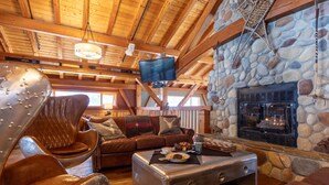 A cozy cabin interior with a stone fireplace, leather furniture, and a television mounted on the ceiling. - Heather Mountain Lodge (Golden)